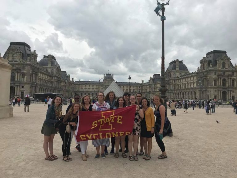 A group of students with a red Iowa State flag holding it facing the camera and smiling in front of a building in France