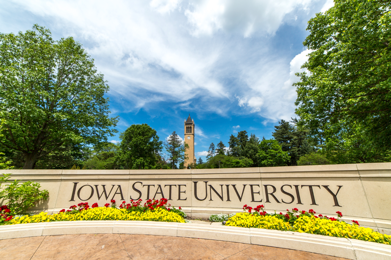 Picture of campus with a blue sky and campanile with a wall that says "Iowa State University" on it