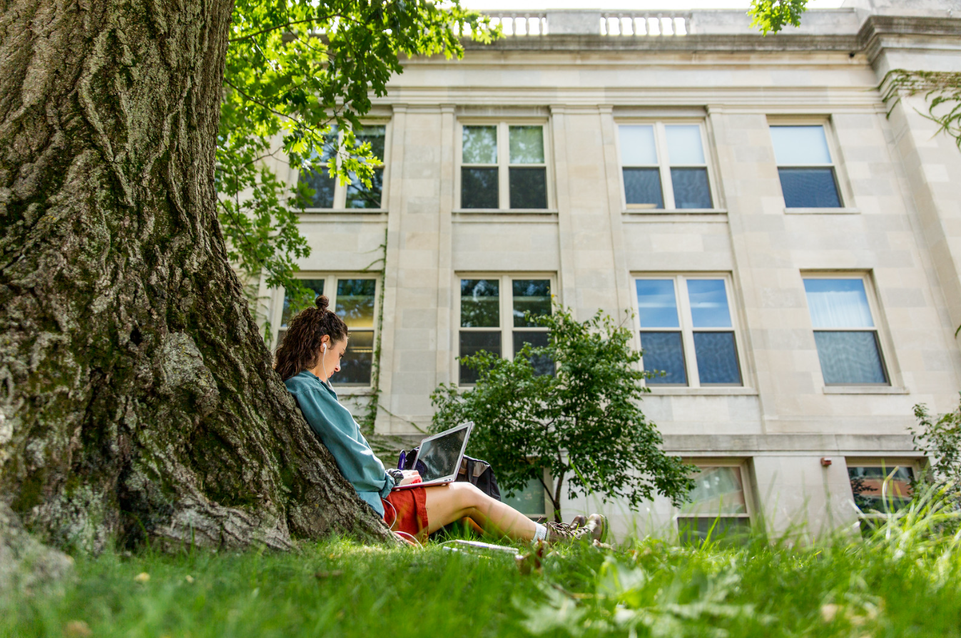 Student sitting with her back against a tree on a laptop with a large, light limestone building in the background. The woman has dark hair, a denim shirt and red shorts. The laptop is resting on her outstretched legs.