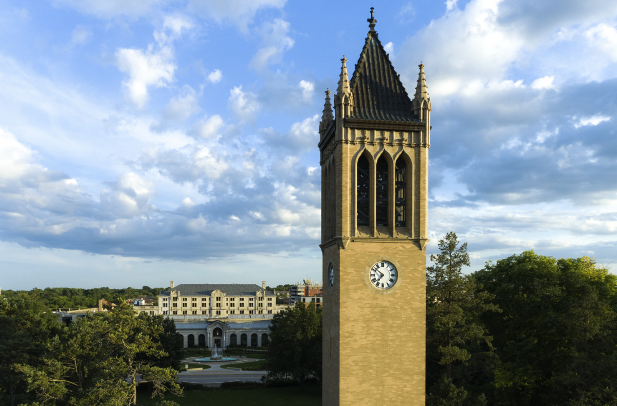 A blue sky with white puffy clouds is in the background of a campanile - a light brick rectangular clock tower.