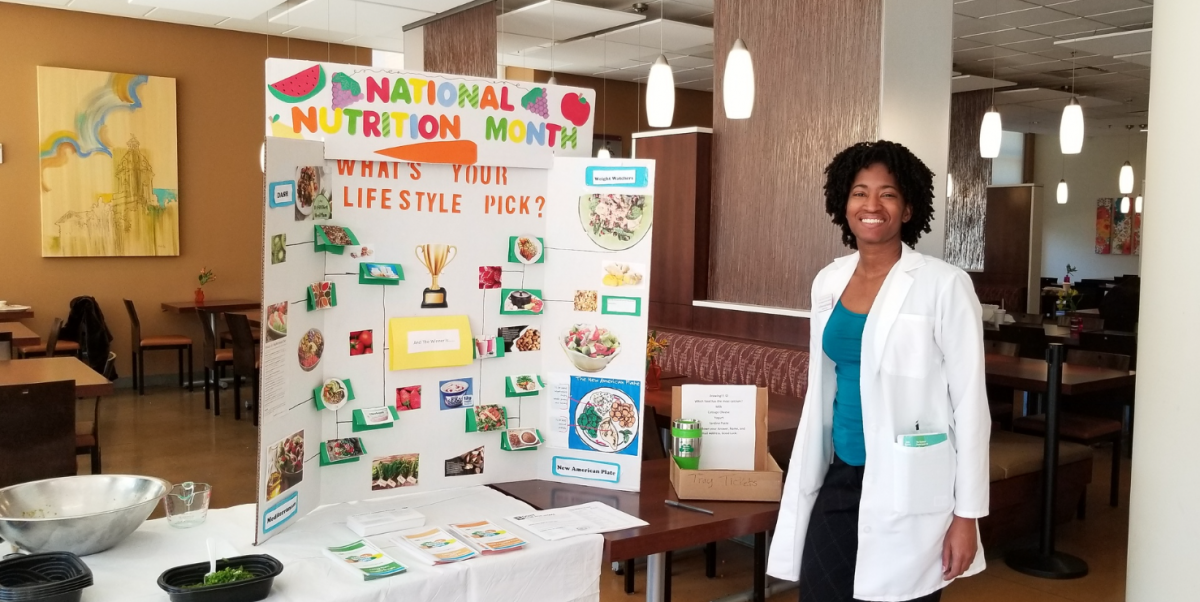 Woman in white lab coat next to a display table that says "National Nutrition Month" on it.